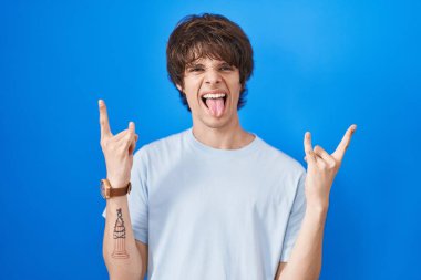 Mavi arka planda dikilmiş çılgın bir ifadeyle bağıran İspanyol genç adam elleri havada rock sembolü yapıyor. Müzik yıldızı. ağır müzik konsepti. 