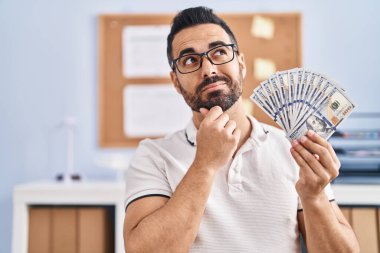 Sakallı ve gözlüklü İspanyol bir genç. Elinde dolar banknotları var. Ciddi bir yüz ifadesi var. 