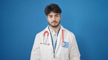 İzole edilmiş mavi arka planda ciddi bir ifadeyle duran genç İspanyol doktor.
