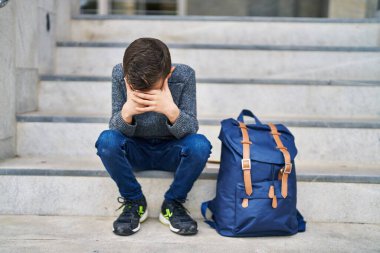 Okulda merdivenlerde oturan endişeli yüzlü sarışın çocuk.