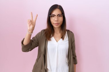 Orta yaşlı Çinli kadın pembe arka planda gözlük takıyor. İki numaralı parmaklarıyla işaret ediyor ve gülümsüyor. Kendinden emin ve mutlu.. 