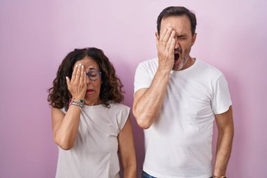 Orta yaşlı İspanyol çift birlikte pembe arka planda esniyorlar. Yarı yüzü, göz ve ağzı elle kapatmaktan yoruluyorlar. Yüzüm acıyor.. 