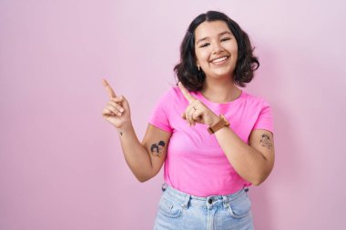 Young hispanic woman standing over pink background smiling and looking at the camera pointing with two hands and fingers to the side. 