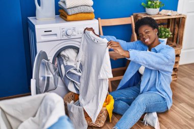 African american woman holding t shirt using washing machine at laundry room