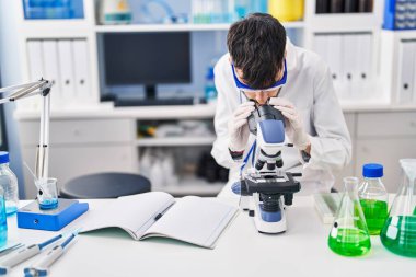Young hispanic man scientist using microscope working at laboratory