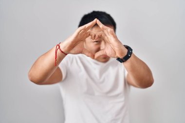 Beyaz arka planda duran genç Çinli adam kalp şeklinde elleriyle ve parmaklarıyla tabelanın içinden bakıp gülümsüyor. 