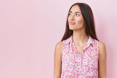 Young hispanic woman with long hair standing over pink background smiling looking to the side and staring away thinking. 