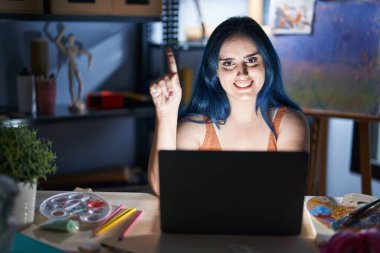 Mavi saçlı genç bir kız, geceleri dizüstü bilgisayarla sanat stüdyosunda oturuyor. Bir numaralı parmağını gösterip onu işaret ederken, kendinden emin ve mutlu bir şekilde gülümsüyor.. 