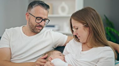 Anne, baba ve bebek emziren aile evdeki kanepede oturuyor.