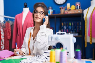 Young beautiful hispanic woman tailor talking on smartphone writing on notebook at clothing factory