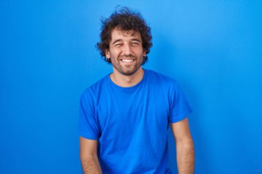 Latin asıllı genç adam mavi arka planda duruyor. Seksi bir ifadeyle kameraya bakıyor. Neşeli ve mutlu bir yüzü var.. 
