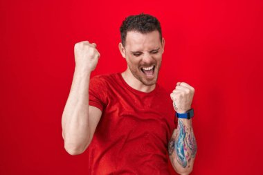 Young hispanic man standing over red background celebrating surprised and amazed for success with arms raised and eyes closed. winner concept. 