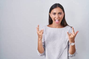 Young hispanic woman standing over white background shouting with crazy expression doing rock symbol with hands up. music star. heavy music concept. 