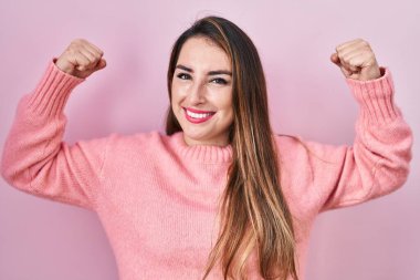 Young hispanic woman standing over pink background showing arms muscles smiling proud. fitness concept. 