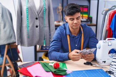 Young hispanic man tailor smiling confident holding dollars at clothing factory
