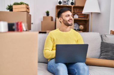 Young hispanic man using laptop sitting on sofa at new home