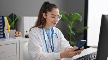 Young beautiful hispanic woman doctor using smartphone working at clinic