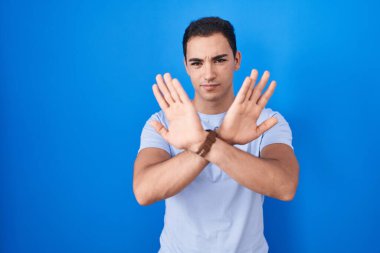 Mavi arka planda duran Latin asıllı genç adam. Reddedilme ifadesi. Kollar ve avuç içleri kesişiyor. 