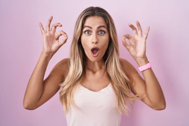 Young blonde woman standing over pink background looking surprised and shocked doing ok approval symbol with fingers. crazy expression 