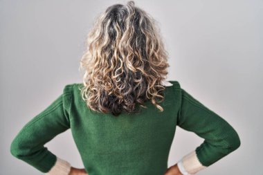 Orta yaşlı bir kadın beyaz arka planda dikiliyor geriye bakıyor kolları vücudunda. 