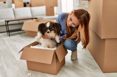 Beyaz bir kadın yeni evinde elinde karton kutunun köpeğiyle yerde oturuyor.