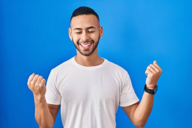 Young hispanic man standing over blue background celebrating surprised and amazed for success with arms raised and eyes closed. winner concept. 