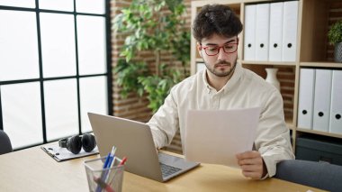 Young hispanic man business worker using laptop reading document at office