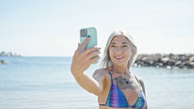 Orta yaşlı, gri saçlı, bikini giyen bir turist kumsalda akıllı telefondan selfie çekiyor.