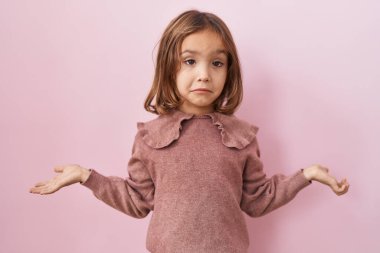 Pembe arka planda duran küçük İspanyol kız elleri ve kolları havada, kafası karışmış bir ifade takınıyor. şüphe kavramı. 