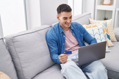 Young hispanic man using laptop and credit card sitting on sofa at home