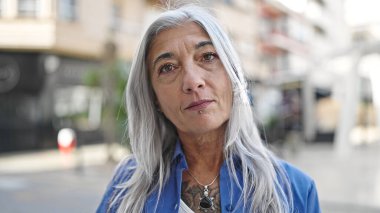 Sokakta ciddi bir ifadeyle duran orta yaşlı, kır saçlı bir kadın.