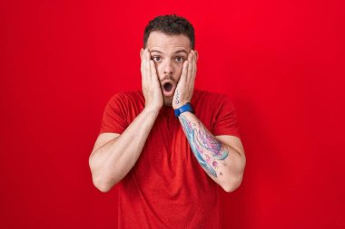 Young hispanic man standing over red background afraid and shocked, surprise and amazed expression with hands on face 