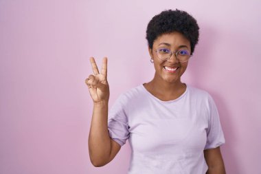 Genç Afro-Amerikalı kadın pembe arka planda duruyor ve iki numaralı parmaklarıyla işaret ederken kendinden emin ve mutlu gülümsüyor.. 