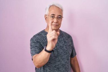 Pembe arka planda duran gri saçlı orta yaşlı bir adam parmağını kaldırmış ve kızgın bir ifadeyle işaret ediyor. 