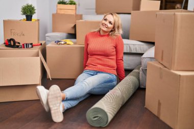 Genç sarışın kadın yeni evinde yerde kendinden emin bir şekilde gülümsüyor.