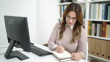 Young beautiful hispanic woman student using computer writing on notebook at library university