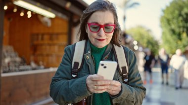 Sokak pazarında akıllı telefon kullanarak sırt çantası takan gri saçlı, olgun İspanyol bir kadın.