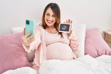 Genç hamile kadın, yatak odasında ekografi tutarak akıllı telefondan selfie çekiyor.