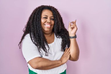 Artı, pembe arka planda duran, yüzünde kocaman bir gülümseme olan, elini kaldırıp kameraya bakan İspanyol bir kadın.. 