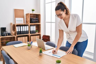 Young beautiful hispanic woman business worker writing on document standing at office