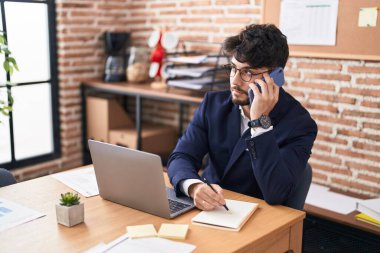 Young hispanic man business worker talking on smartphone write on notebook at office