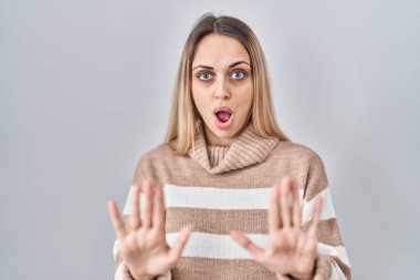 İzole edilmiş arka planda balıkçı yaka kazak giyen genç sarışın bir kadın. Avuçlarını kaldırıyor, reddediyor ve iğrenç bir ifadeyle reddediyor. Dur ve yasakla. 