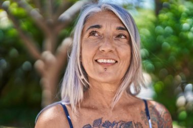 Orta yaşlı, kır saçlı, gülümseyen, kendine güvenen bir kadın parkın kenarına bakıyor.