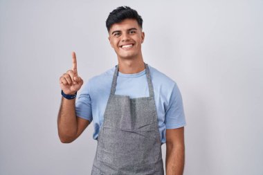 Beyaz arka planda önlük giyen İspanyol genç adam bir numaralı parmağını gösterip işaret ederken kendinden emin ve mutlu bir şekilde gülümsüyor.. 