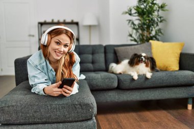 Genç beyaz kadın evde köpekle kanepede müzik dinliyor.