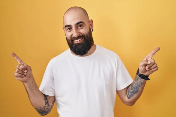 髭と入れ墨を持つ若いヒスパニック系の男は 異なる方向に指で自信を持って指して笑顔黄色の背景に立っている 広告のコピースペース — ストック写真