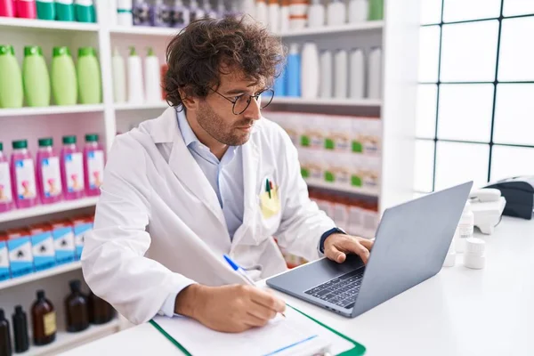 Young Hispanic Man Pharmacist Writing Document Using Laptop Pharmacy —  Fotos de Stock