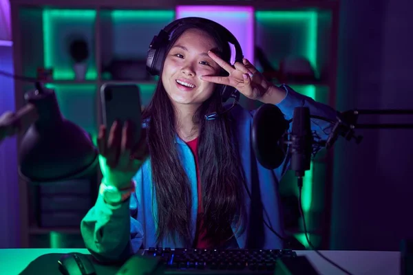 若いアジアの女性は スマートフォンでビデオゲームをプレイ顔の上に指で平和のシンボルをやって 明るい勝利を示す笑顔 — ストック写真