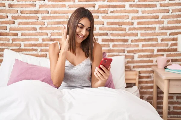 笑顔で勝利を祝う自宅でベッドの上に座ってスマートフォンを使用して若いヒスパニック系の女性と手を上げて勝者の式 — ストック写真