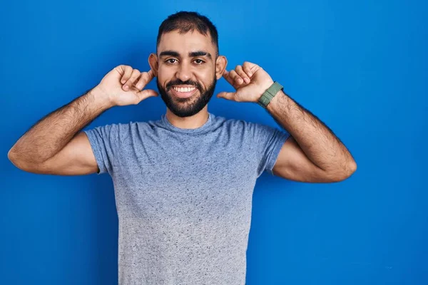 中東の男性のひげの青い背景に立って指で耳を引く笑みを浮かべて 面白いジェスチャー オーディション問題 — ストック写真